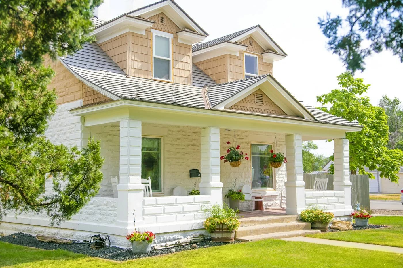 A two-storey white and warm-tan family house with a wide covered front porch, white columns and hanging flower baskets, on a sunny day